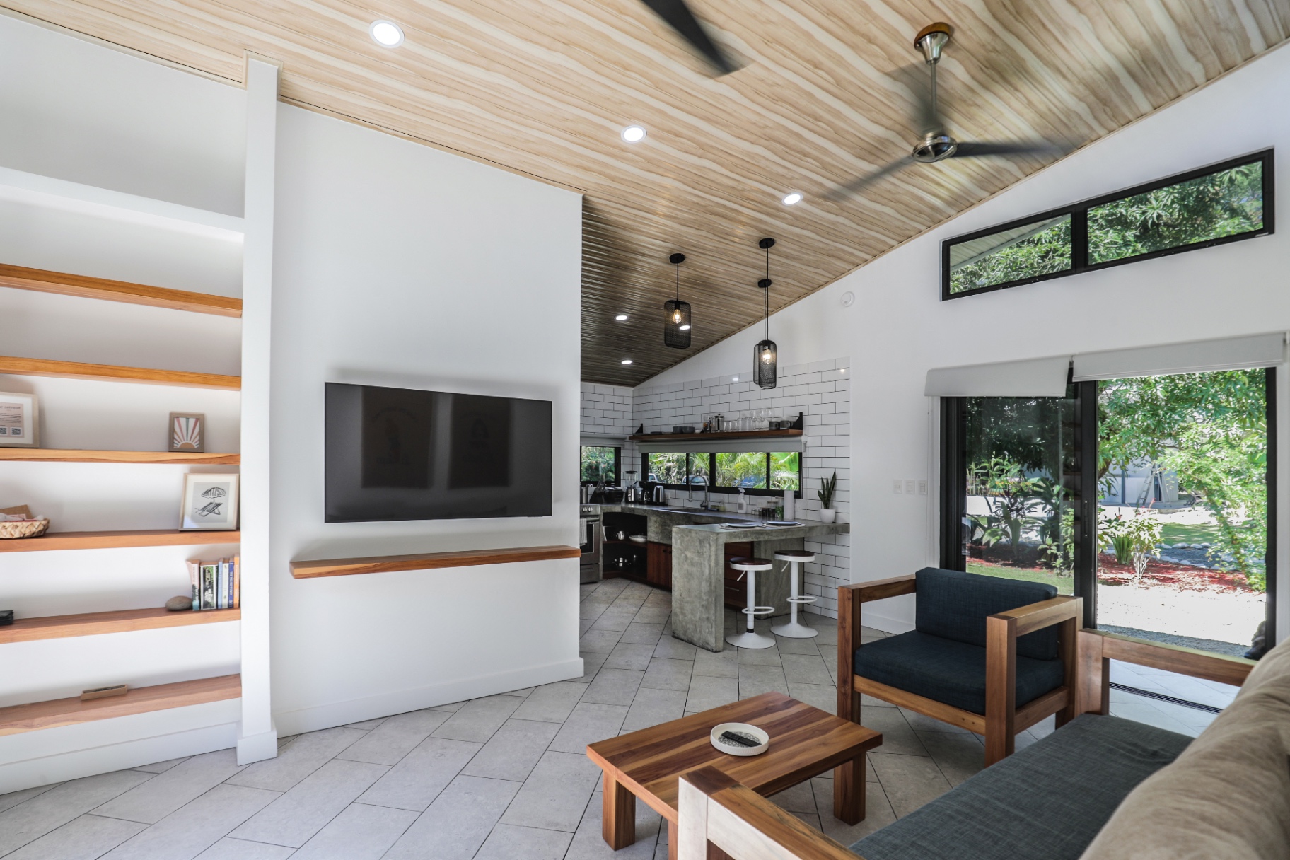 Open-concept living room with wall-mounted TV, teak shelves and view into the full kitchen in Unit 1