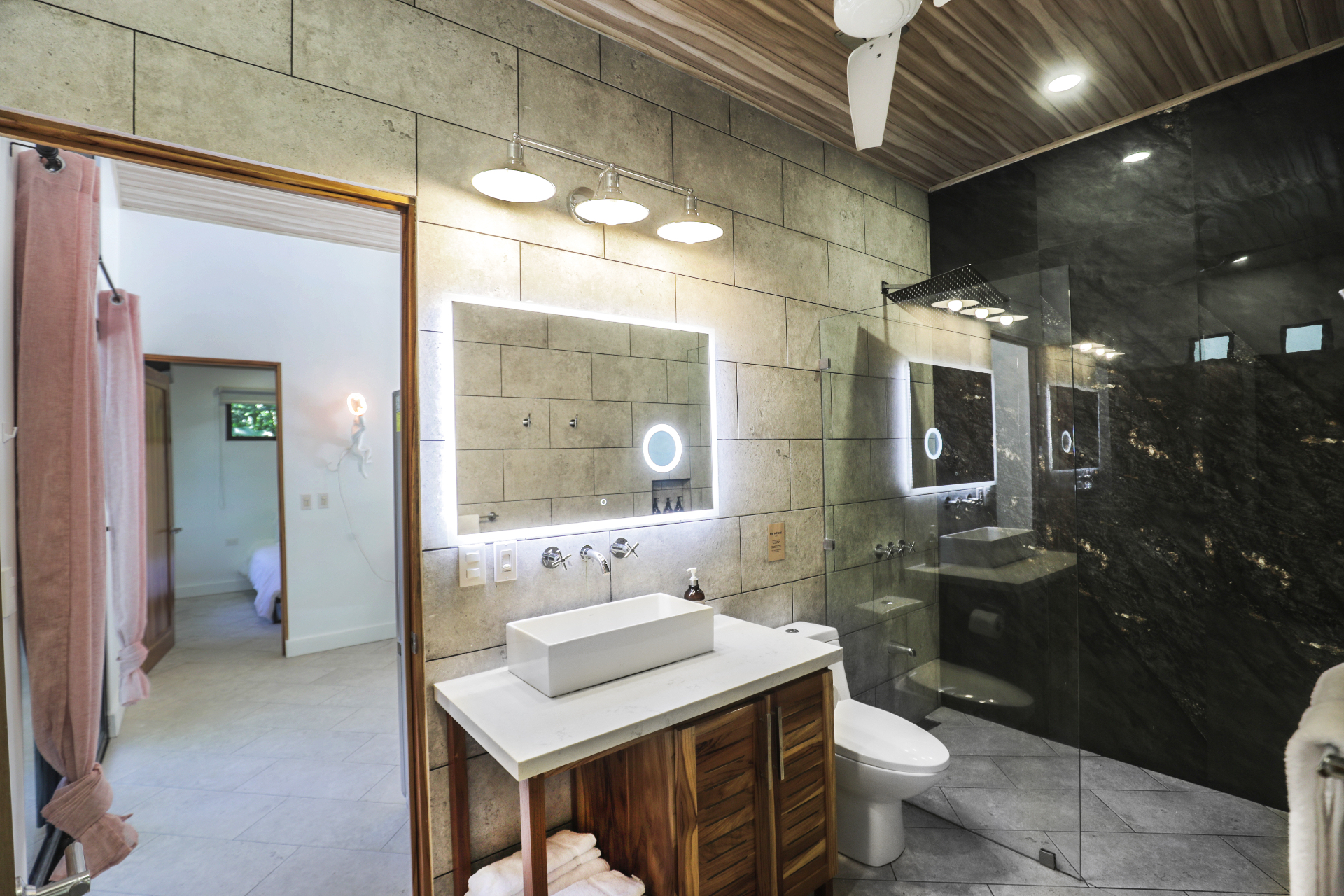 Ensuite bathroom with concrete-tile walls, backlit mirror, wood vanity and black stone rain shower in Unit 4